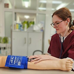 Eine Studierende bei einer Übung zum Blutdruckmessen. Auf dem Tisch lieg ein Übungsarm, angelegt eine Druckmanschette.