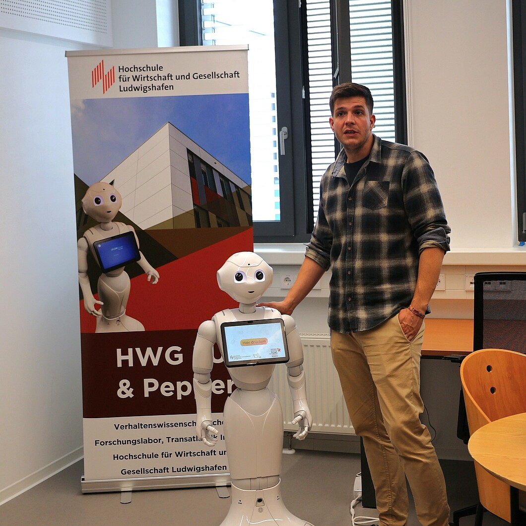 Ein Mann steht neben dem Roboter Pepper und zeigt auf dessen Bildschirm. Der Roboter steht vor einem Banner, und die Präsentation findet in einem Raum statt, der auf die Institution im Hintergrund hinweist.