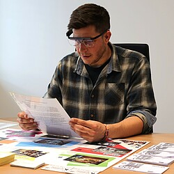 A man wears eye-tracking glasses and reads a document while participating in a business game that simulates a visit to a shopping center. In front of him on the table, various stores and areas of the shopping center are depicted on a play area