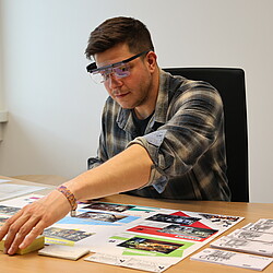 A man wears eye-tracking glasses and reaches for a pile of notes during a business game that simulates a visit to a shopping center. Various store areas and play money are spread out on the table in front of him.