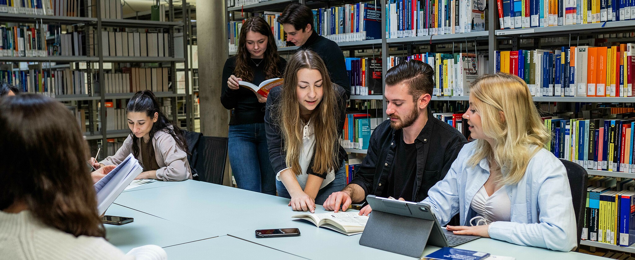 A group of students in the library, with three students sitting in the foreground and studying together.