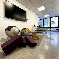 View of the simulation circle room, in the foreground you can see various cushions, a birthing stool, an exercise ball and a beanbag. A hospital bed can be seen in the background.