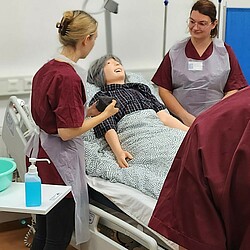 Students doing an exercise at a hospital bed, you can see two students with gowns at the bed. A manikin is lying in the bed.