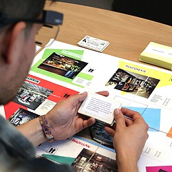 Ein Mann trägt eine Eyetracking-Brille und betrachtet eine Karte, während er an einem Planspiel teilnimmt, das den Besuch eines Einkaufszentrums simuliert. Verschiedene Geschäfte und Bereiche des Einkaufszentrums sind auf der Spielfläche abgebildet.