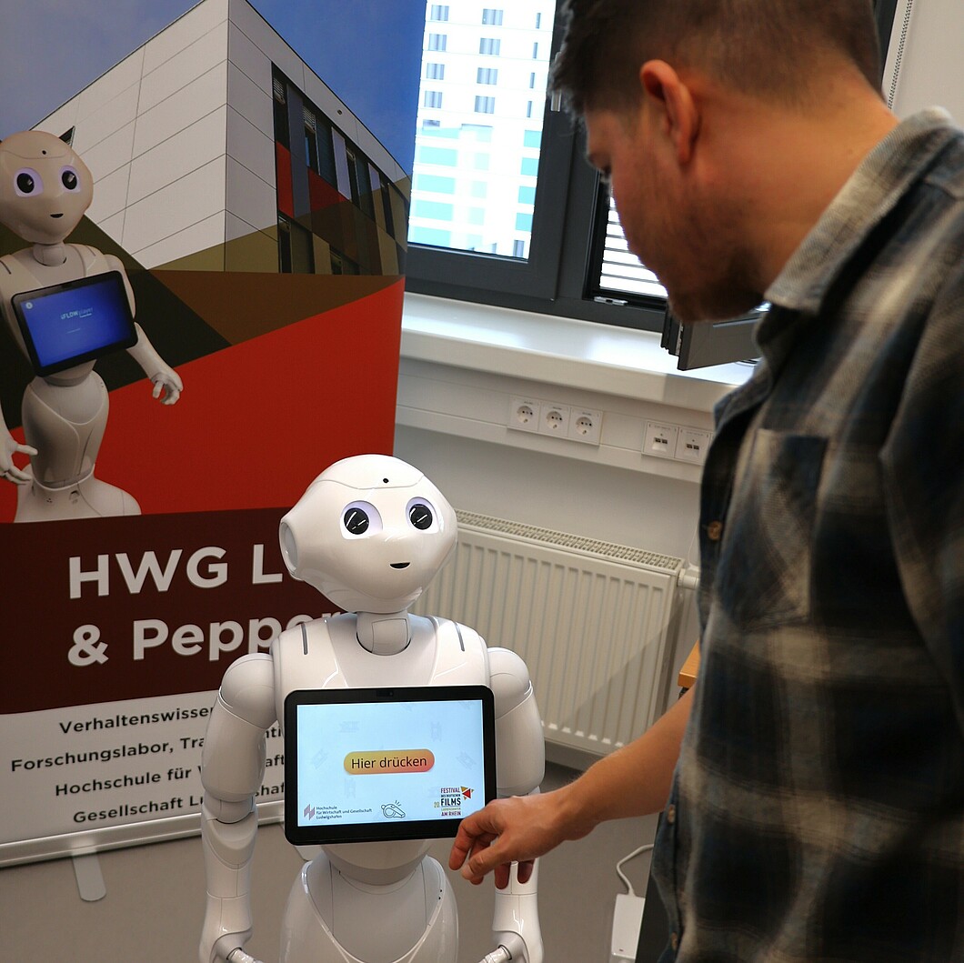 Ein Mann interagiert mit dem Roboter Pepper, während dieser eine Nachricht auf seinem Bildschirm zeigt. Der Roboter steht vor einem Banner, das ihn und eine Institution im Hintergrund zeigt.