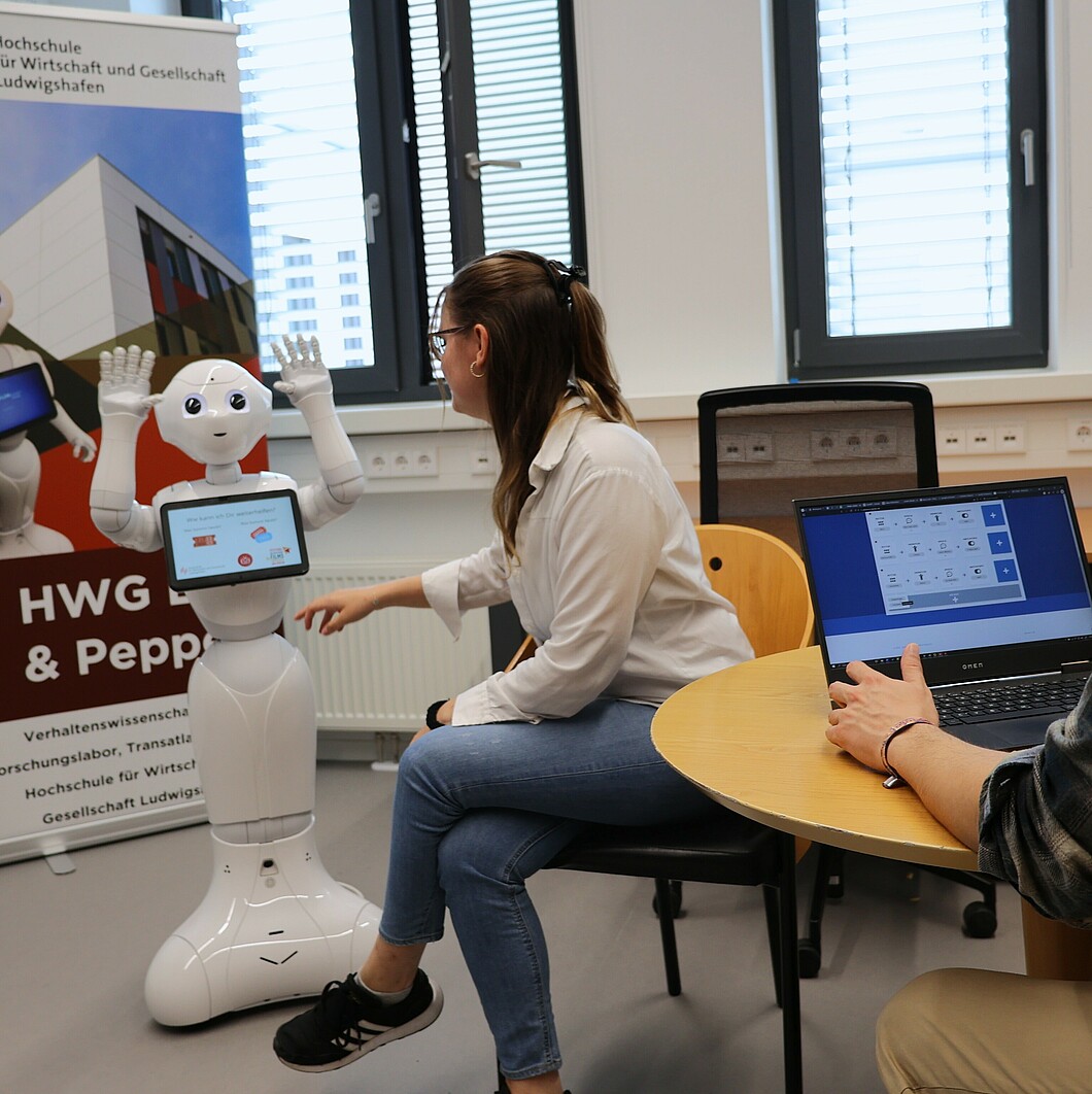 Der Roboter Pepper hebt seine Hände, während eine Frau mit ihm interagiert und ein Mann im Hintergrund sitzt. Der Roboter scheint eine Geste zu machen, die Teil seiner Demonstration ist.