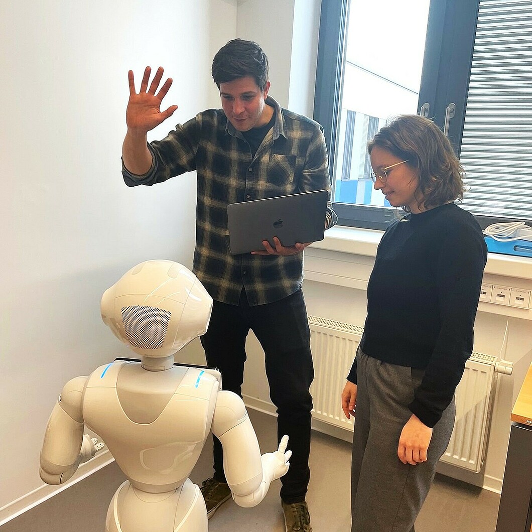 Ein humanoider Roboter Pepper interagiert mit zwei Personen in einem Raum. Ein Mann hebt die Hand zur Begrüßung des Roboters, während eine Frau interessiert zuschaut.