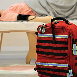 A staged scene, with a red emergency kit in the foreground and a manikin lying in bed in the background.