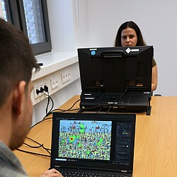 A man looks at an eye-tracking heat map on a laptop while a woman sits in front of an eye-tracking device in the background. The image shows the analysis of gaze behavior in a complex visual scenario.