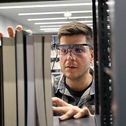 A person wears eye-tracking glasses and looks intently at a row of books on a shelf in a library.