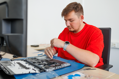 A person (trainee) is working with a screwdriver on an open laptop on a blue surface. Next to it is a monitor on a desk in a bright office space.