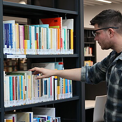 Ein Mann trägt eine Eyetracking-Brille, während er in einer Bibliothek nach einem Buch greift. Der Versuch untersucht sein Blickverhalten bei der Auswahl von Büchern aus dem Regal.