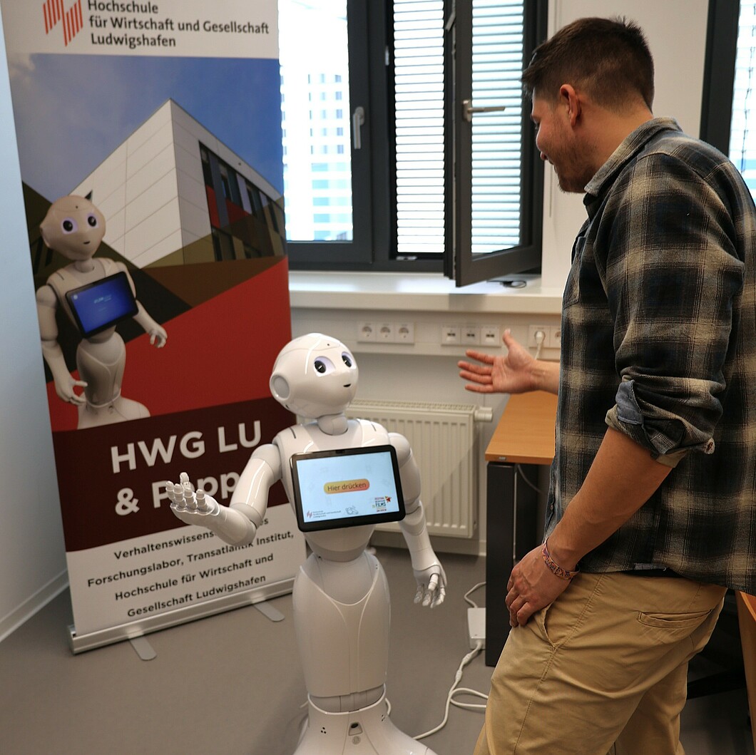 Ein Mann spricht mit dem Roboter Pepper, während dieser seine Hände hebt. Der Roboter zeigt auf seinem Touchscreen eine Nachricht an, und der Mann steht in einer aktiven Interaktion mit ihm