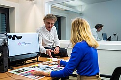 Ein Mann sitzt auf einem Tisch und erklärt einer Frau verschiedene Materialien. Auf dem Bildschirm ist das Logo der Hochschule für Wirtschaft und Gesellschaft Ludwigshafen zu sehen.