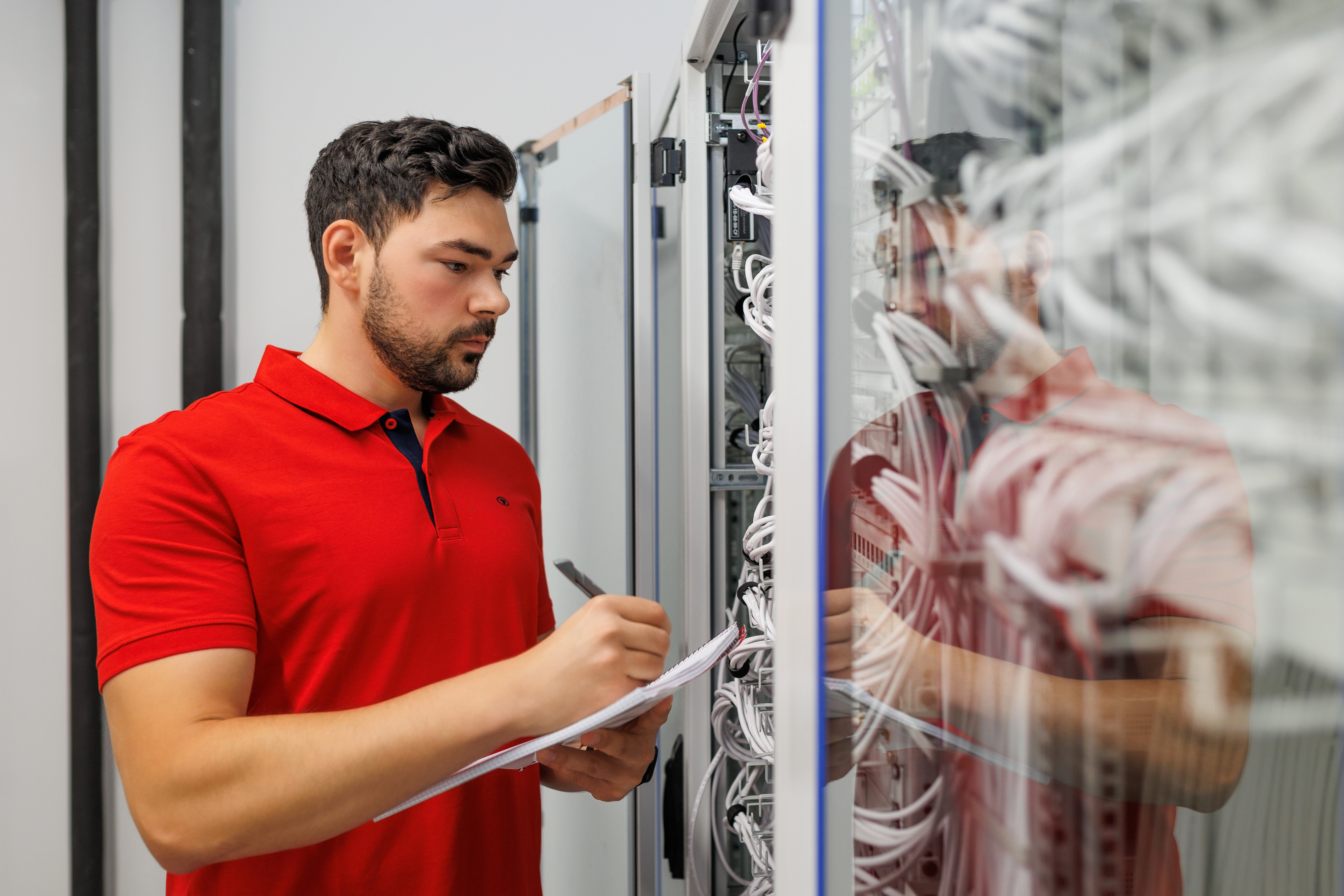 A person (trainee) is standing at the open network cabinet and writing something down on a clipboard. Many white patch cables can be seen in the cabinets, the person is reflected in the glass door.